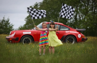 Two young girls waving flags in front of retro car in excitement for Retro Cannonball