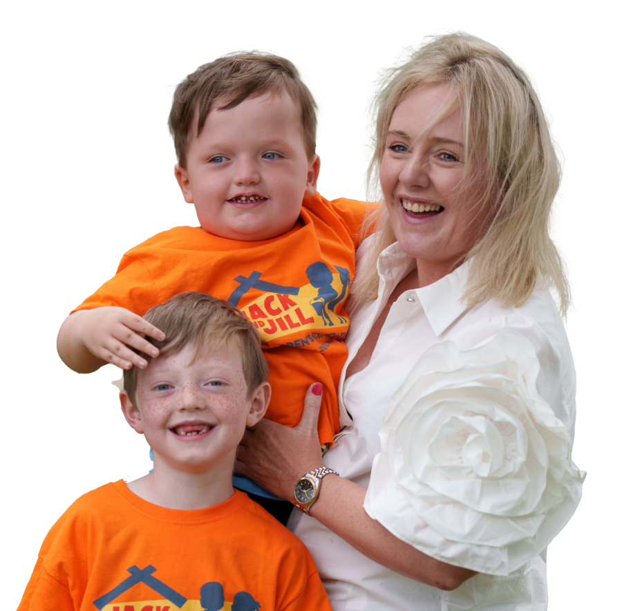 A smiling blonde woman wearing a white collared shirt holds a young boy wearing a bright orange Jack and Jill Children's Foundation t-shirt. Another young boy with freckles smiles in front of them while the boy being held rests his hand gently on his head. Set against a plain white background.