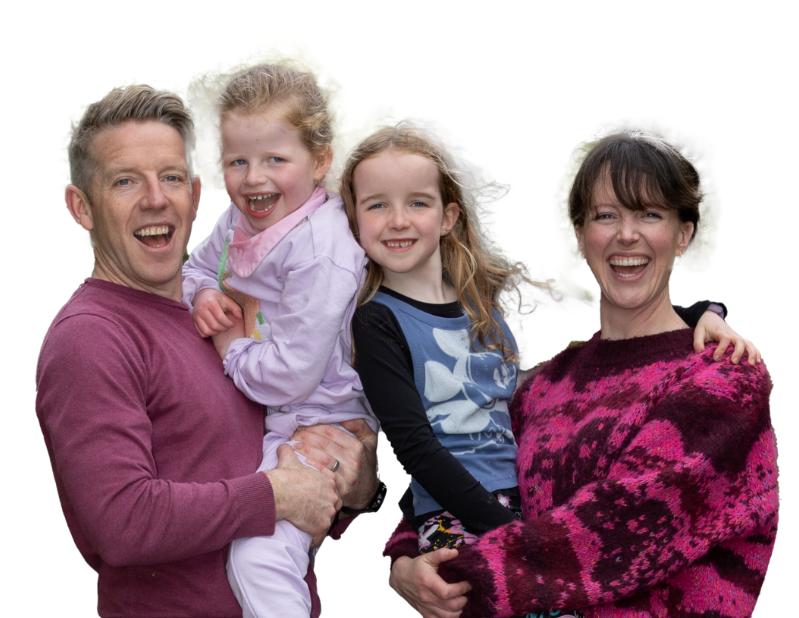 A happy family of four smiling brightly against a plain white background. A man on the left in a maroon sweater holds a young girl wearing a light pink tracksuit. On the right, a woman in a vibrant pink and purple patterned sweater holds a young girl with long brown hair wearing a blue graphic shirt and colorful patterned leggings.