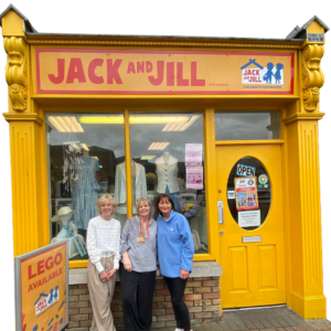 Three women smiling outside the bright yellow storefront of the Jack and Jill Children's Foundation charity boutique in Newbridge, with a display window showcasing clothing and a sidewalk sign that reads LEGO AVAILABLE