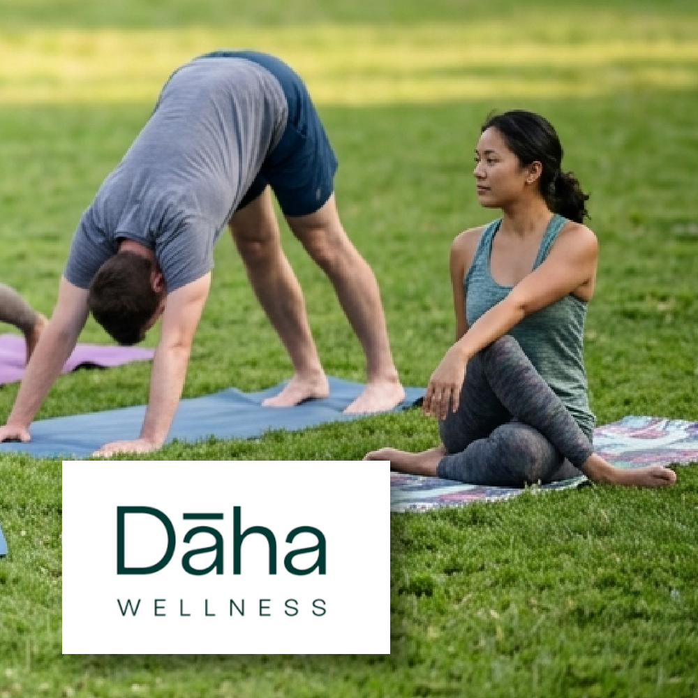 Dāha Wellness A woman sitting cross-legged in a spinal twist and a man performing a downward-facing dog pose on yoga mats outdoors on a grassy lawn, with the "Dāha WELLNESS" logo overlaid in the lower-left corner.