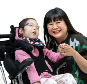 Mila, a young girl with additional needs in a supportive wheelchair, smiling and holding hands with her mother