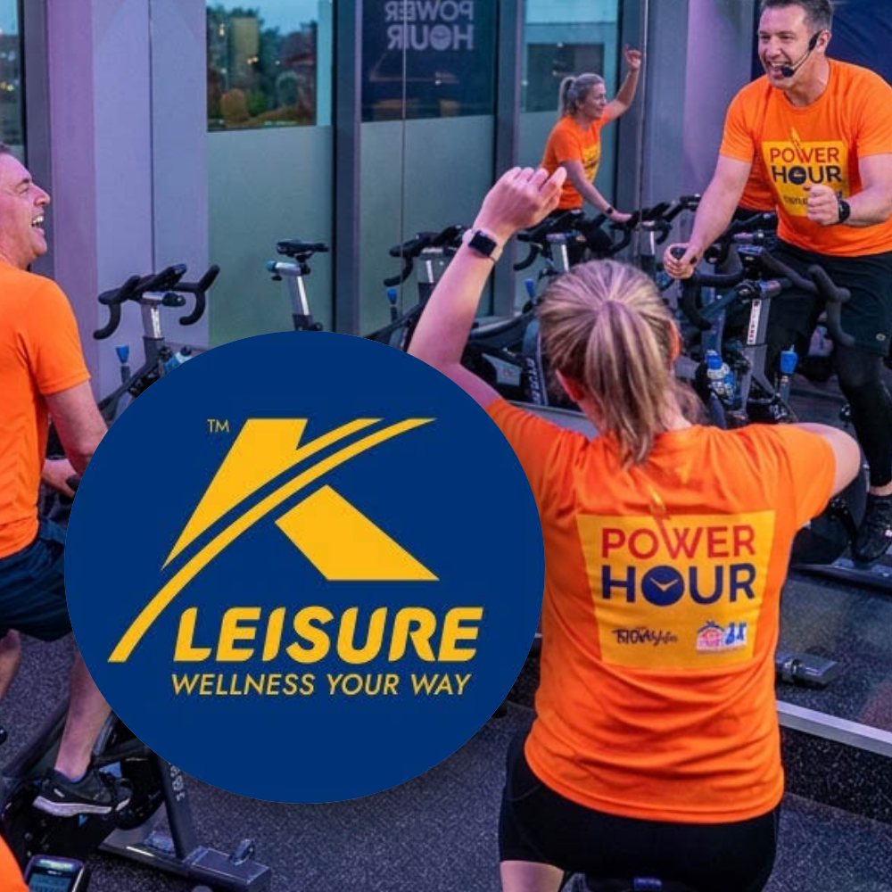 K-Lesiure Spinathon A group of people in orange "Power Hour" shirts participating in a spin class at a gym, with a circular blue logo overlay displaying the text "K Leisure Wellness Your Way".