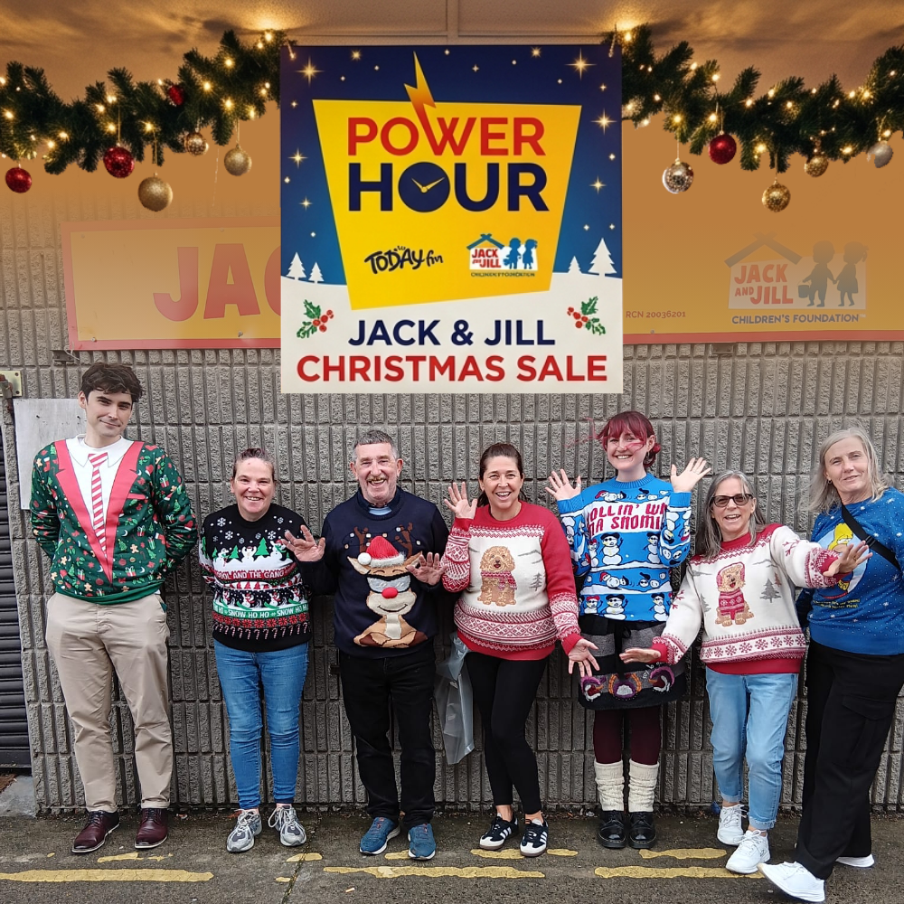 Power Hour Artane Seven people wearing festive Christmas jumpers waving outside a store, beneath a holiday garland and a Jack and Jill Christmas Sale Power Hour sign.