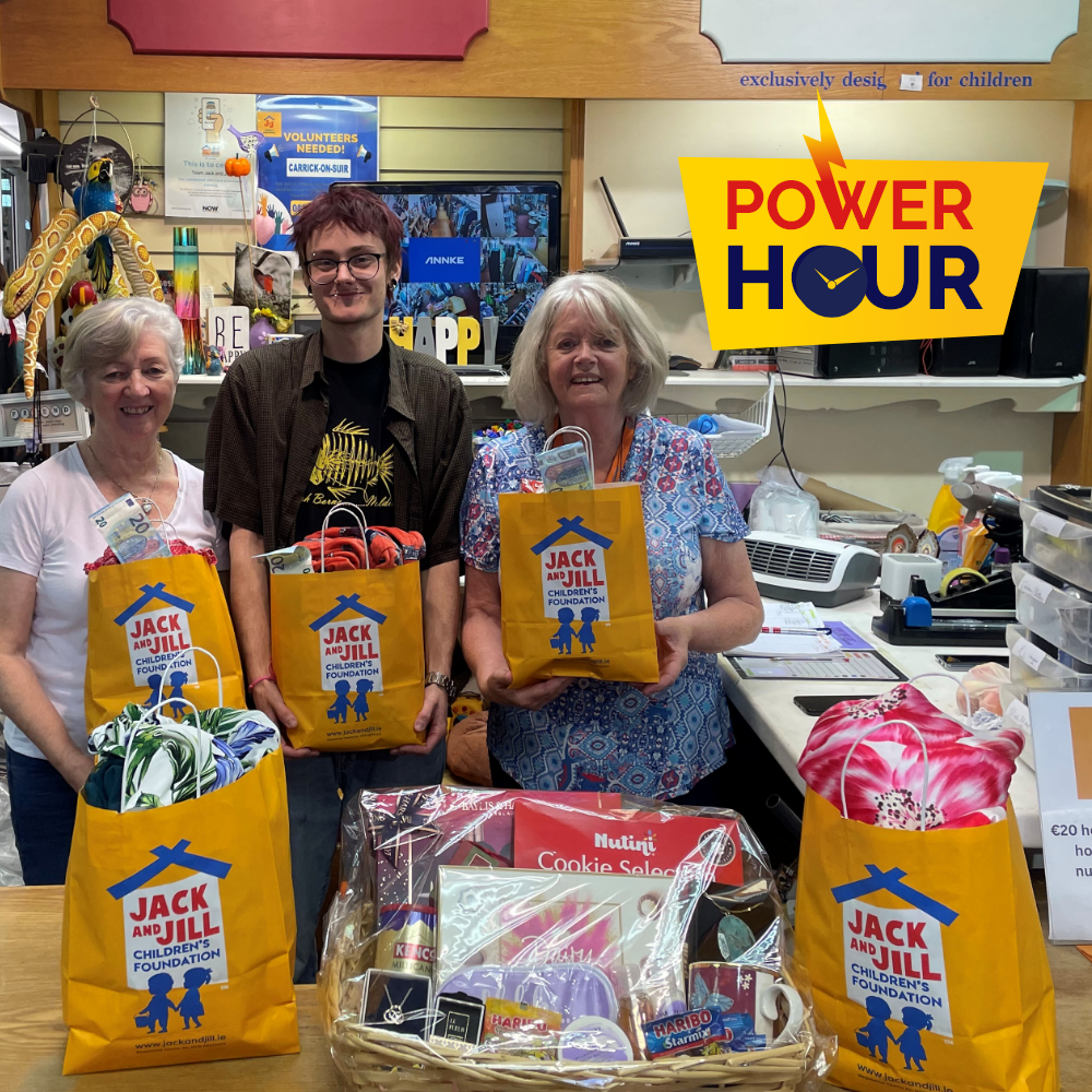 Power Hour Carrick on Suir 2 Three people behind a store counter displaying yellow Jack and Jill Children's Foundation gift bags filled with cash and items, alongside a gift hamper, with a yellow Power Hour logo in the top right corner.