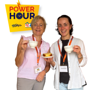 Two smiling women wearing lanyard badges holding teacups and a slice of cake, next to a yellow "Power Hour" logo featuring Today FM and the Jack and Jill Children's Foundation.