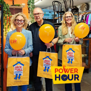 Three Jack and Jill charity boutique team members holding orange balloons and yellow branded shopping bags inside the store, featuring the Power Hour logo.