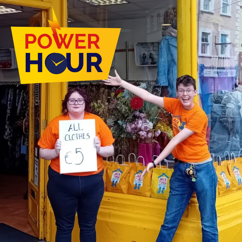 Power Hour youghal Two Jack and Jill charity boutique staff members in orange t-shirts standing outside a store window holding an "All Clothes €5" sign, featuring the Power Hour logo.