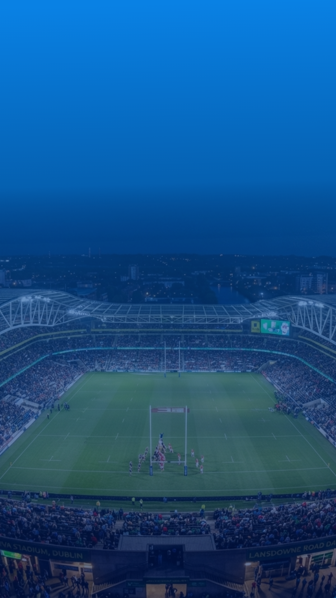 Aerial view of a packed rugby stadium at night, with the illuminated pitch and goalposts visible beneath a blue overlay.