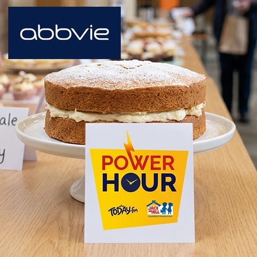 bake sale AbbVie Victoria sponge cake dusted with powdered sugar on a cake stand next to a 'Power Hour' Today FM and Jack and Jill Foundation sign, with an AbbVie logo in the top corner.