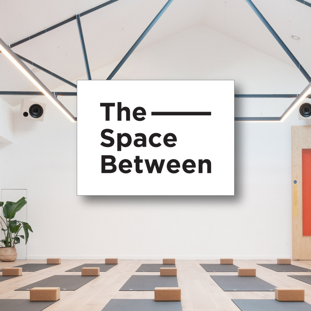 the space between An empty yoga studio with wooden flooring, grey yoga mats, and cork blocks laid out in rows, featuring a prominent rectangular graphic in the center with the black text "The Space Between".