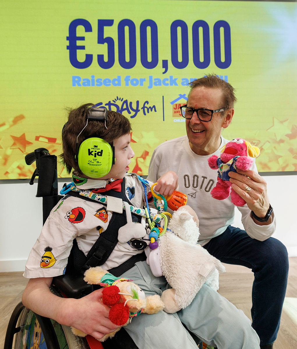 Ian Dempsey and James Garrahan 007 Broadcaster Ian Dempsey smiling and holding a puppet toy while interacting with James in a wheelchair, with a background banner celebrating €500,000 raised for Jack and Jill.