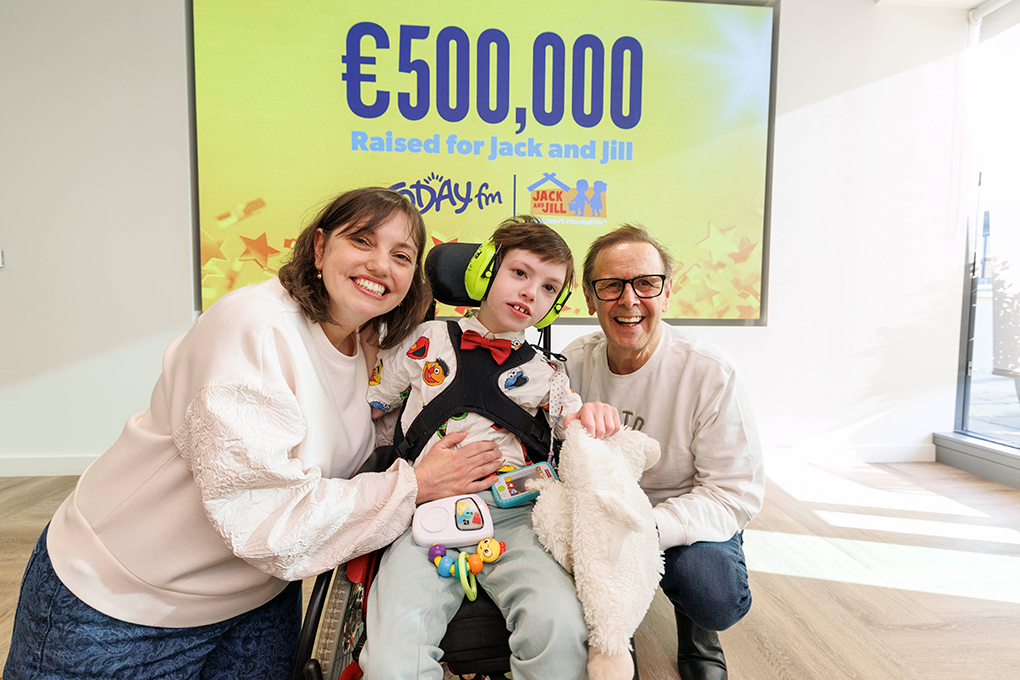 Ian Dempsey and the Garrahan Family 009 James, a family member, and Ian Dempsey smiling together in front of a screen displaying '€500,000 Raised for Jack and Jill'.
